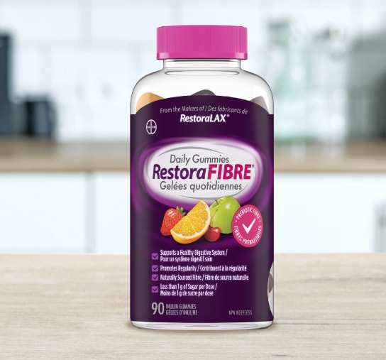 Un flacon transparent de gelées quotidiennes RestoraFIBRE® avec un capuchon rose et une étiquette violette montrant des images de fruits et des affirmations relatives à la santé est posé sur une surface en bois clair dans une cuisine floue.