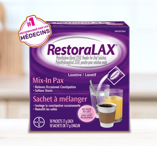 Une boîte violette de sachets à mélanger RestoraLAX® est posée sur une surface en bois, avec un bol de fruits et légumes verts et un arrière-plan flou.